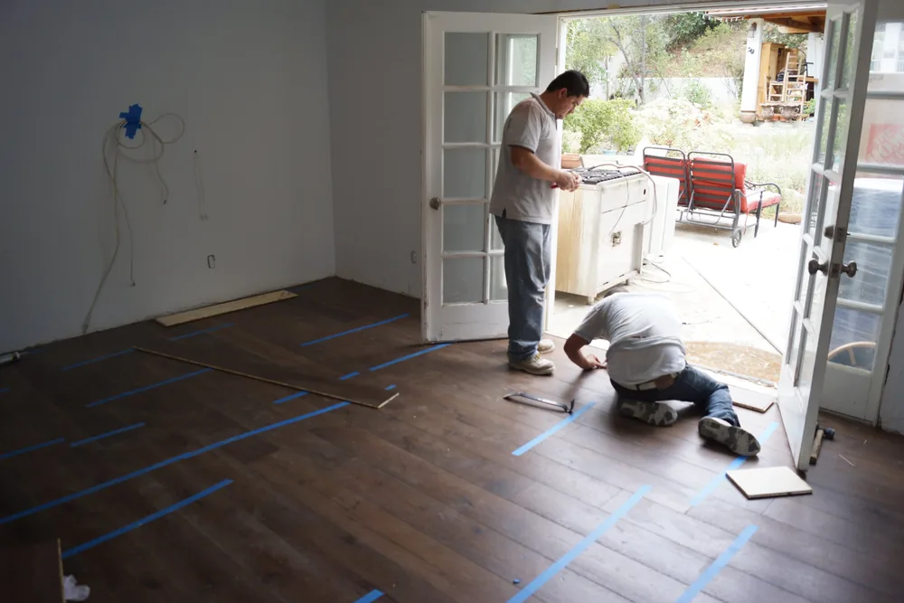 Installers laying dark wide-plank hardwood flooring near French doors in a Pasadena home